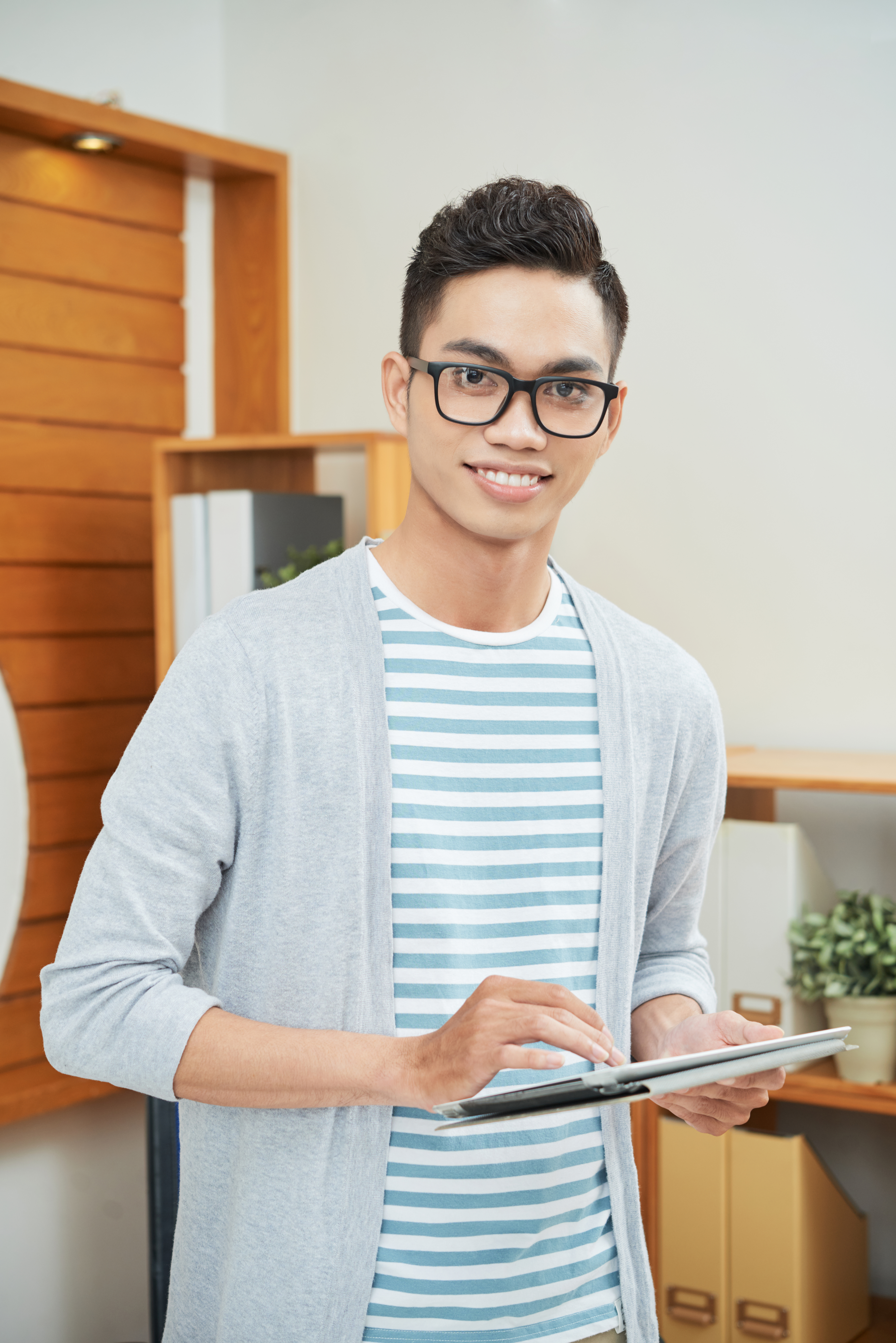 Casual young ethnic man in eyeglasses smiling while standing with touchpad in office